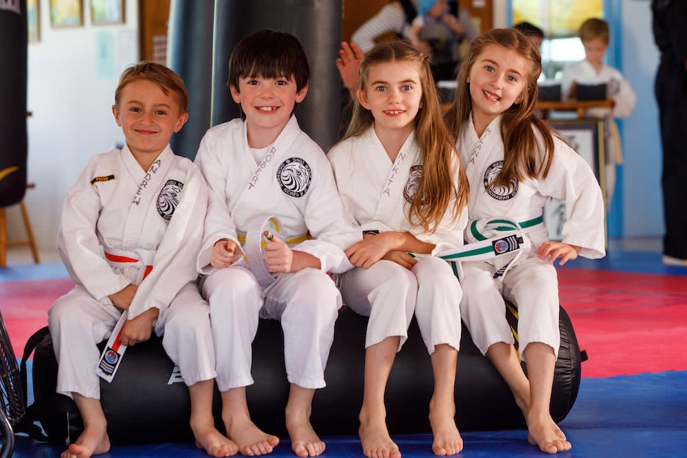 Kids in karate uniforms smiling and sitting together on a mat at Polander Academy in Bethesda, showcasing a friendly and supportive martial arts environment.