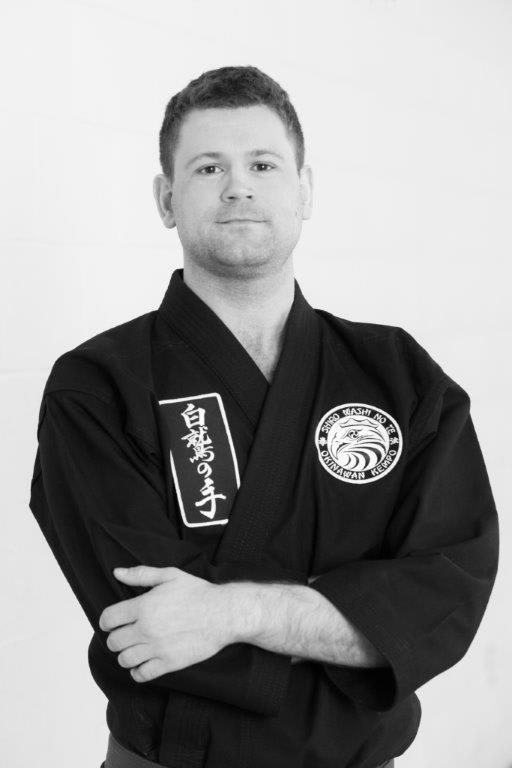 Matthew Cowie, Assistant Instructor at Polander Academy of Martial Arts, wearing a black karate gi with a logo, arms crossed, in a studio setting in Bethesda.